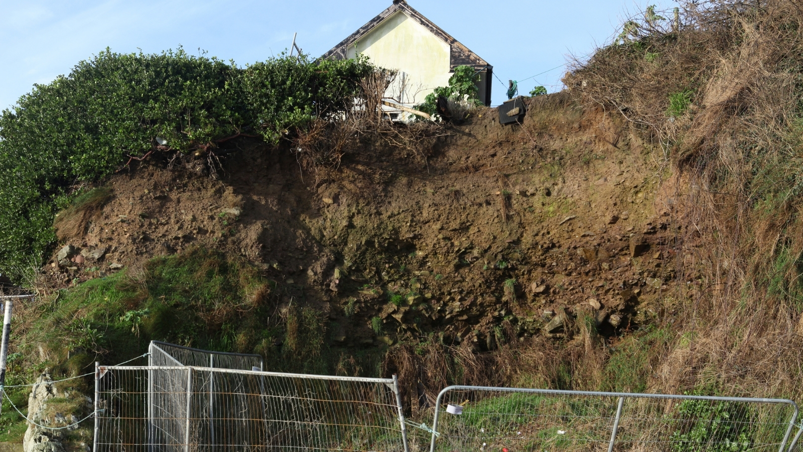A house perched above a collapsed cliff face, with temporary fencing at the foot of the slope.