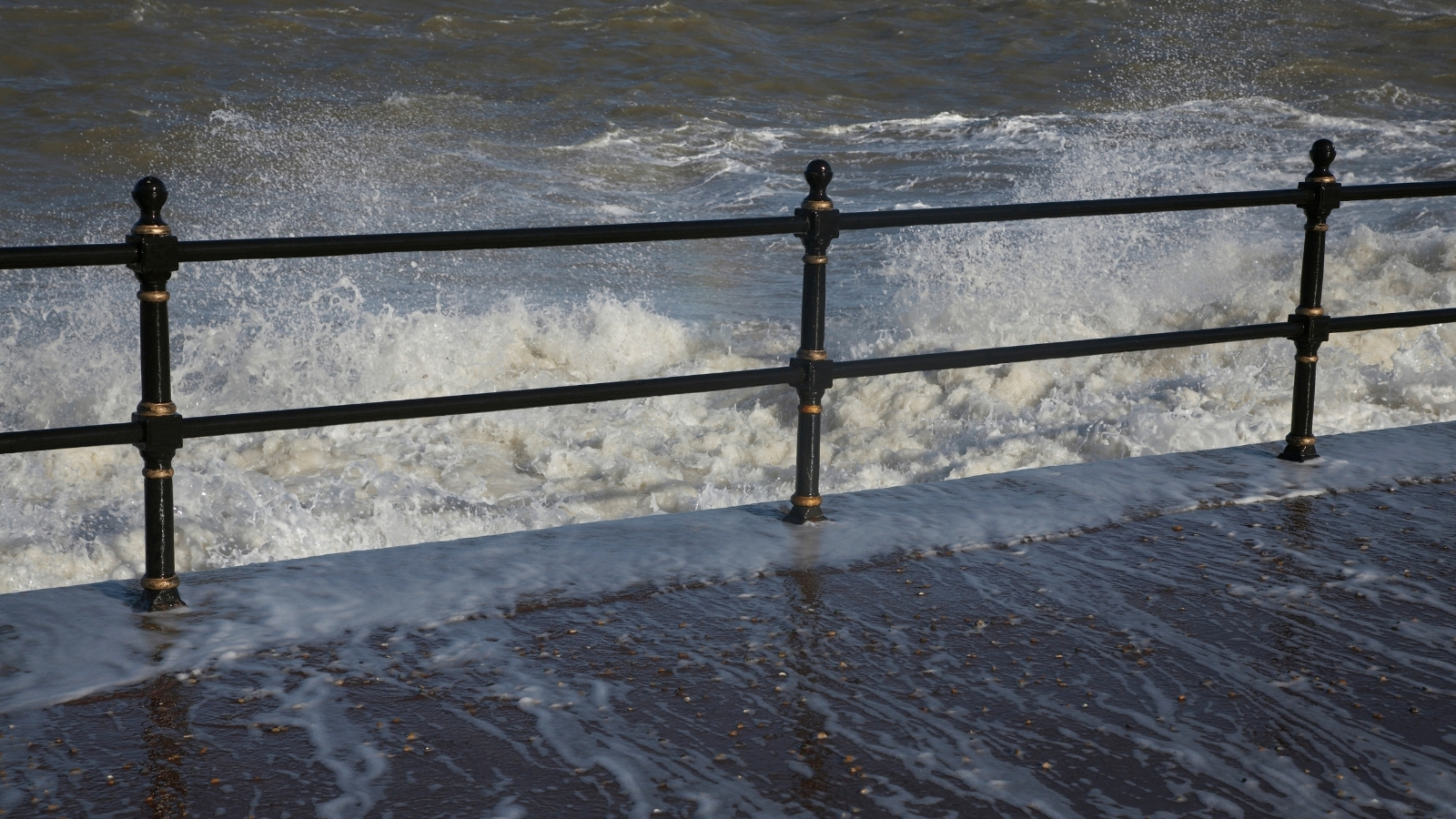 Waves overtopping a promenade railing and washing across the wet pavement.